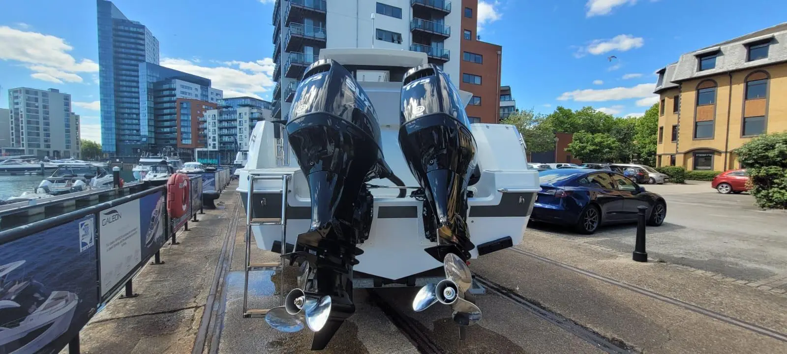 A sleek powerboat with twin outboard motors is docked near modern buildings and parked cars in a marina setting.